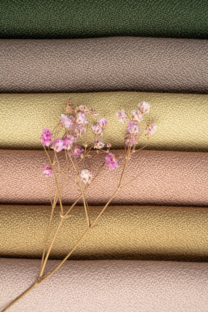A sprig of small pink flowers lies on top of stacked, textured fabric in muted green, beige, taupe, and gold—discover the best materials for acoustic panels and how they optimize sound absorption for various spaces.