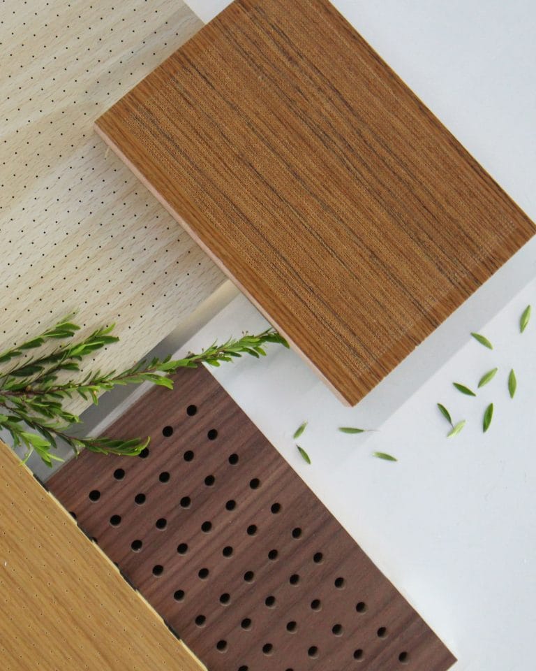 Three rectangular wood panels with different finishes and perforations are arranged on a white surface, next to a small green branch and scattered leaves—discover the best materials for acoustic panels and how they optimize sound absorption for various spaces.