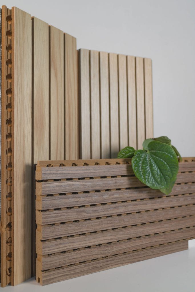Several wooden acoustic wall panels with vertical grooves stand upright on a white surface. Featuring fire-safe timber cladding that doesn’t sacrifice craft, a green leafy plant adds a touch of nature to the display.