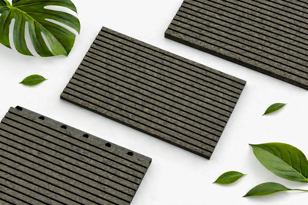 Three rectangular, black, grooved tiles are arranged on a white background, surrounded by several green leaves, including a large monstera leaf and a few smaller leaves, creating a modern and natural composition.