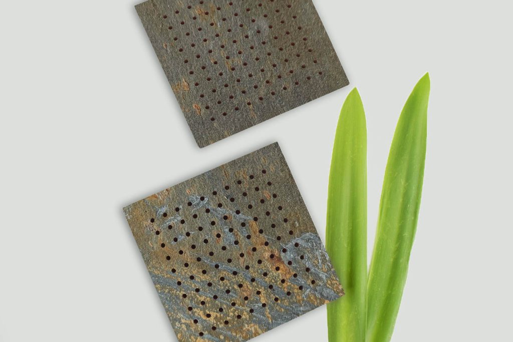 Two square, perforated pieces of dark, textured material are arranged diagonally above two green leaves on a light background.