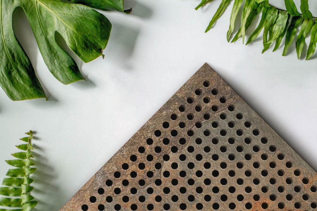 A rusty perforated metal sheet with round holes is positioned in the bottom right corner, surrounded by green monstera and fern leaves on a white background.