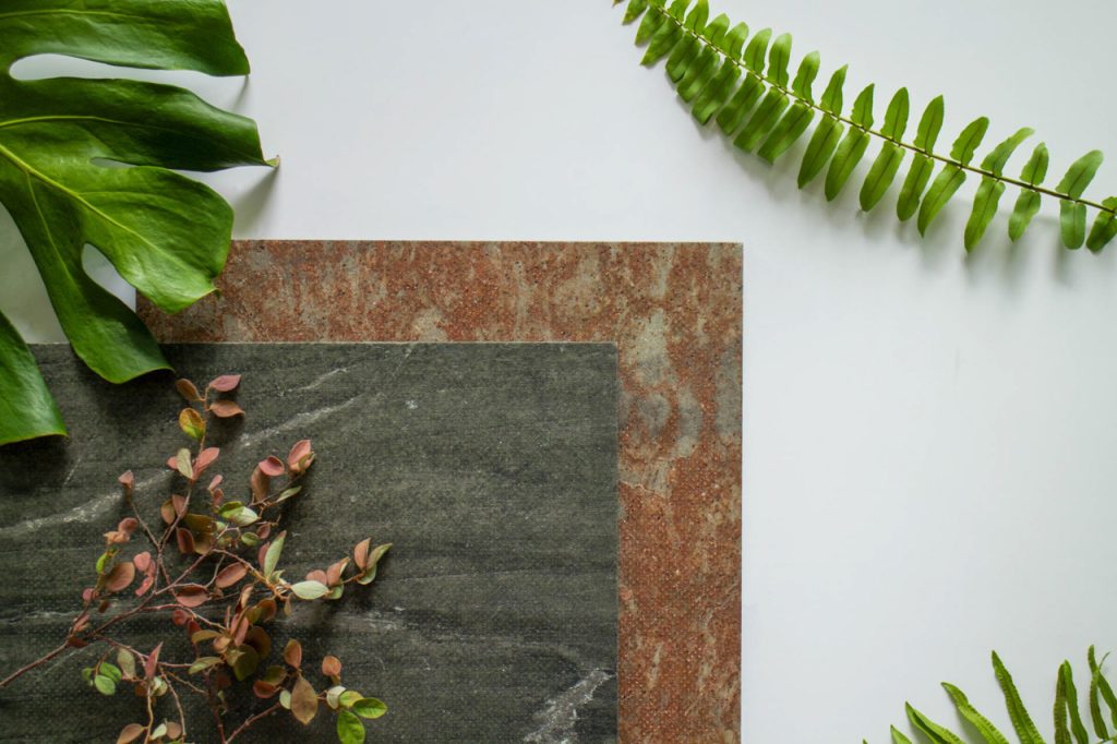 Close-up of micro perforated wood panels with finely drilled holes, arranged on a light surface and accented with a few green leaves. The panels display varied wood tones and distinct grain patterns.