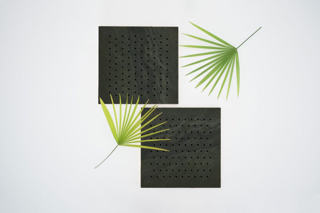 Two square, black, perforated panels are placed on a white background, with two green palm leaves arranged diagonally around them. The scene has a minimalistic, modern look.