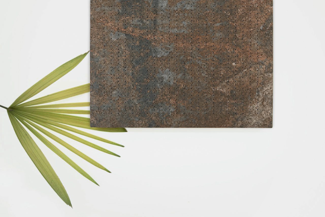 A green palm leaf rests on a white surface beside a square, rust-colored, perforated metal sheet with weathered texture.