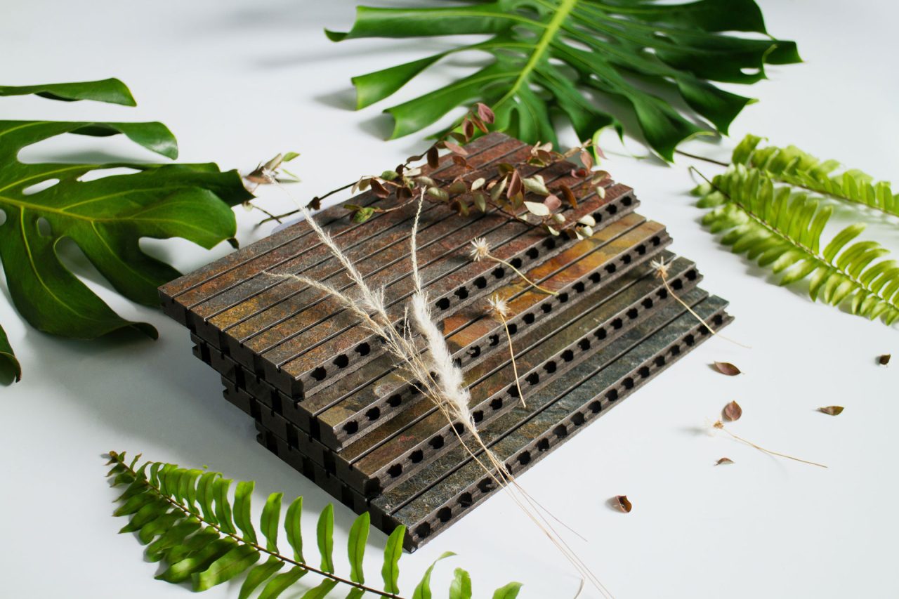 A stack of rectangular metal bars with holes sits on a white surface, surrounded by various green leaves and dried plants.