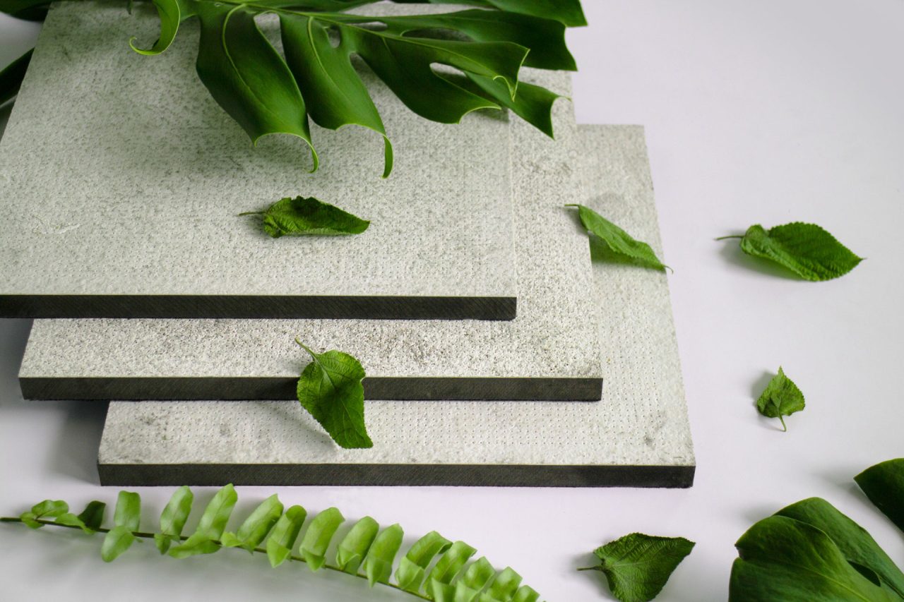 Three textured stone or concrete slabs are stacked on a white surface, surrounded by various green leaves and foliage, creating a natural and minimalist aesthetic.