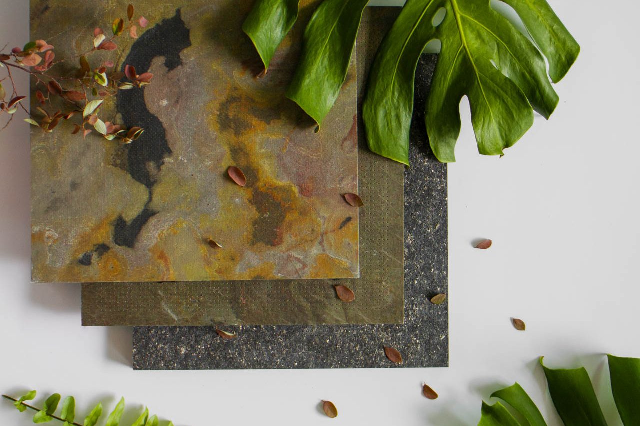 Three square stone tiles with earthy colors are arranged on a white surface, surrounded by large green monstera leaves, a fern, and scattered brown seeds.